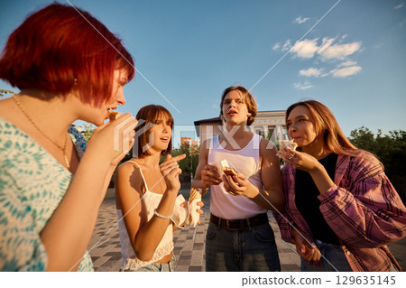 Friends standing outdoors in warm light eating snacks and talking together 129635145