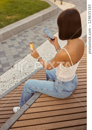 Woman sitting on bench taking a photo of hotdog using smartphone outdoors 129635165