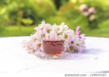 Summer still life; Transparent cup of black tea and bunch of pink cosmos flowers on a white tablecloth in a blooming summer garden by sunny day 129635587