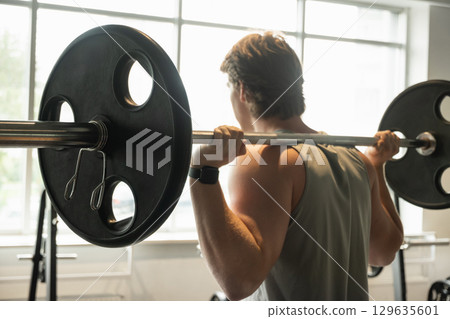 Young caucasian man or bodybuilder exercising with barbell at fitness center. Cross training. Male athlete lifting weights. Squat bar. Back view. Young caucasian man or bodybuilder exercising with barbell at fitness center. Cross training. Male athlete lifting weights. Squat bar. Back view. 129635601