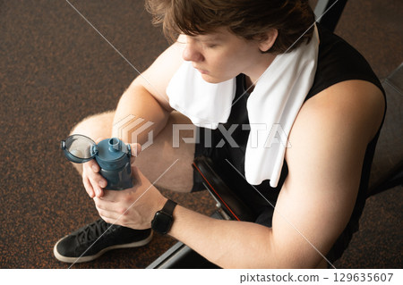 Man sitting in gym holding water bottle and towel on his neck. Moment of relaxation and refreshment after workout. Man sitting in gym holding water bottle and towel on his neck. Moment of relaxation and refreshment after workout. 129635607