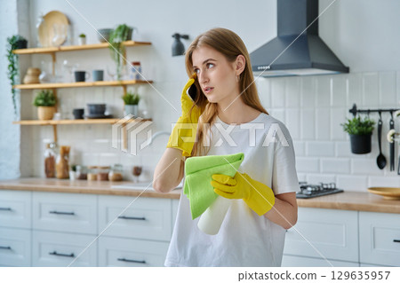 Young woman in gloves with rag cleaning spray talking on phone in kitchen Young woman in gloves with rag cleaning spray talking on phone in kitchen 129635957