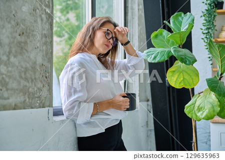 Mature tired, stressed woman standing near window Mature tired, stressed woman standing near window 129635963