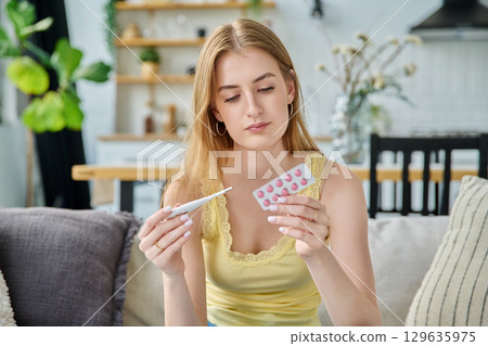 Sad upset teenage girl looking at thermometer holding blister pack of pills sitting at home 129635975