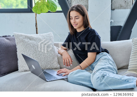 Smiling teenage girl looking at laptop, sitting on sofa at home Smiling teenage girl looking at laptop, sitting on sofa at home 129636992
