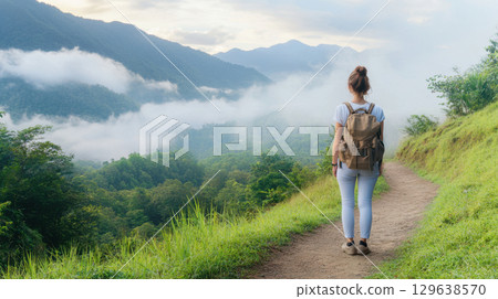 Dreamy summer hiking scene of a young woman with a vintage backpack standing on a winding trail, gazing at misty mountains and lush green wilderness. 129638570