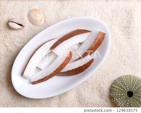 Fresh coconut meat, in oval white bowl, on beach sand with shells. Opaque white flesh of a fresh and young coconut, cut into strips, and ready to eat. Seed of the palm tree Cocos nucifera. Food photo 129638575