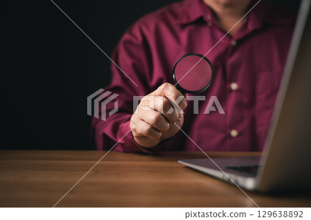 A man holds a magnifying glass near a laptop, symbolizing investigation, research, or scrutiny. The dark background emphasizes analysis, cybersecurity, inspection, and digital security. 129638892