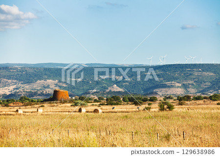 Ancient Nuraghe in Sardinian countryside with hay bales and wind turbines 129638986