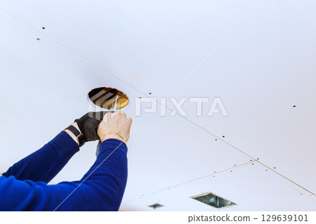 Worker cuts hole in drywall ceiling for lighting installation in residential space during renovation Worker cuts hole in drywall ceiling for lighting installation in residential space during renovation 129639101