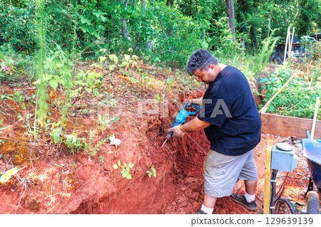 Man digging in red soil, cultivating land, preparing for planting in scenic outdoor area during works daytime Man digging in red soil, cultivating land, preparing for planting in scenic outdoor area during works daytime 129639139