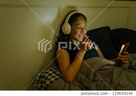A Young Woman Enjoys Listening to Music on Her Smartphone While Relaxing Peacefully in Bed A Young Woman Enjoys Listening to Music on Her Smartphone While Relaxing Peacefully in Bed 129639246
