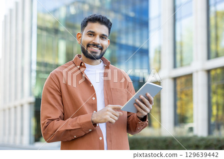 A cheerful man dressed casually stands outside a modern building, holding a tablet. He smiles confidently while using the device, showcasing technology in a professional yet casual setting. A cheerful man dressed casually stands outside a modern building, holding a tablet. He smiles confidently while using the device, showcasing technology in a professional yet casual setting. 129639442