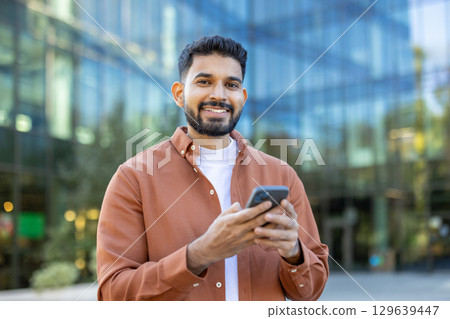 A cheerful man standing outdoors uses his smartphone in a professional urban setting. His happiness and connection to technology exemplify productivity and positivity in a contemporary lifestyle. 129639447
