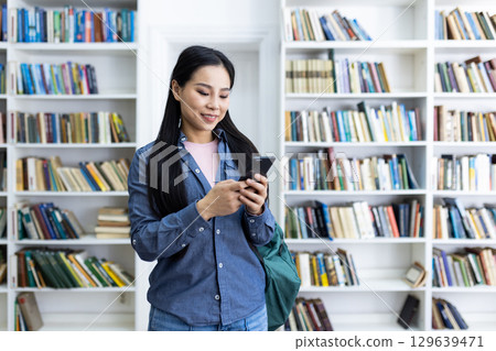 A smiling young woman using her smartphone in a library filled with bookshelves. She appears focused and happy, wearing casual clothing, against the vivid backdrop of neatly arranged books A smiling young woman using her smartphone in a library filled with bookshelves. She appears focused and happy, wearing casual clothing, against the vivid backdrop of neatly arranged books 129639471