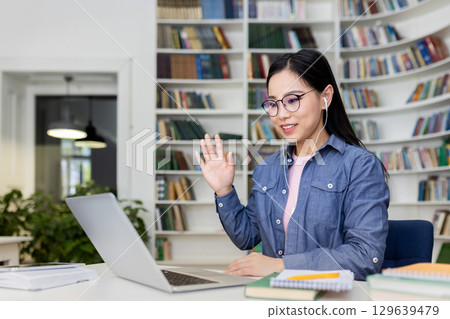 A young woman participates in a virtual meeting using a laptop, sitting in a library. She is wearing headphones, waving, and appears engaged in a lively digital interaction. A young woman participates in a virtual meeting using a laptop, sitting in a library. She is wearing headphones, waving, and appears engaged in a lively digital interaction. 129639479