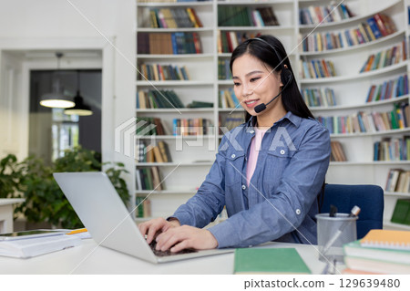 A professional woman sits in a library, wearing a headset, working on her laptop, providing remote support or conducting an online meeting in a focused and organized setting. 129639480