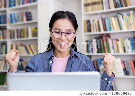 A happy young woman in glasses joyfully celebrates after accomplishing work on her laptop in a library, surrounded by bookshelves, reflecting achievement, success, and motivation. A happy young woman in glasses joyfully celebrates after accomplishing work on her laptop in a library, surrounded by bookshelves, reflecting achievement, success, and motivation. 129639482
