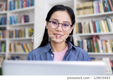A young woman in glasses is sitting in front of her laptop, smiling warmly. The setting appears to be a well-organized library, emphasizing learning, focus, and academic pursuits. A young woman in glasses is sitting in front of her laptop, smiling warmly. The setting appears to be a well-organized library, emphasizing learning, focus, and academic pursuits. 129639483
