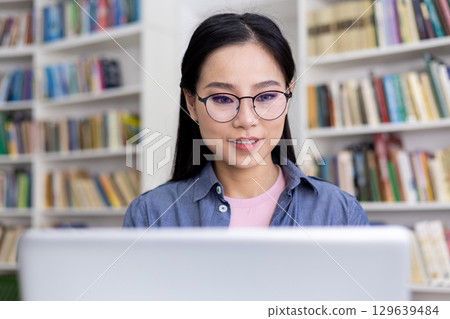 A young woman wearing glasses working on a laptop in a library setting. Shelves filled with books are arranged in the background, creating a calm and intellectual atmosphere while she concentrates. A young woman wearing glasses working on a laptop in a library setting. Shelves filled with books are arranged in the background, creating a calm and intellectual atmosphere while she concentrates. 129639484