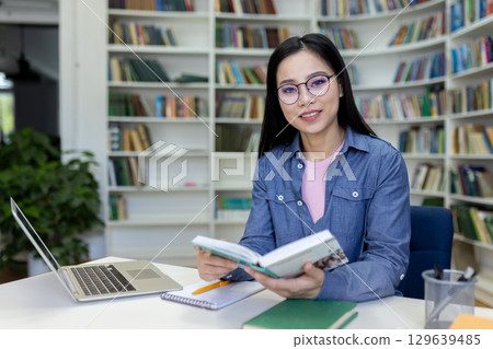 A bright library serves as the backdrop for a young woman reading a book while working at a desk with a laptop, capturing a scene of dedication and learning. A bright library serves as the backdrop for a young woman reading a book while working at a desk with a laptop, capturing a scene of dedication and learning. 129639485