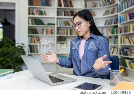 A woman sitting at a desk in a library, gesturing frustratedly while working on her laptop, surrounded by books, expressing problem-solving or challenges in a study or professional context. A woman sitting at a desk in a library, gesturing frustratedly while working on her laptop, surrounded by books, expressing problem-solving or challenges in a study or professional context. 129639486