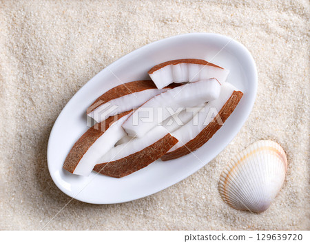 Fresh coconut meat, in oval white bowl, on beach sand with shell. Opaque white flesh of a fresh and young coconut, cut into strips, and ready to eat. Seed of the palm tree Cocos nucifera. Food photo 129639720