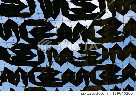 Intriguing Camouflage Netting Overhead, Casting Unique Shadows Against a Clear Blue Sky with Soft Clouds, Creating an Artistic Display of Shape and Color Variation for Enthusiastic Viewer Engagement. 129640200