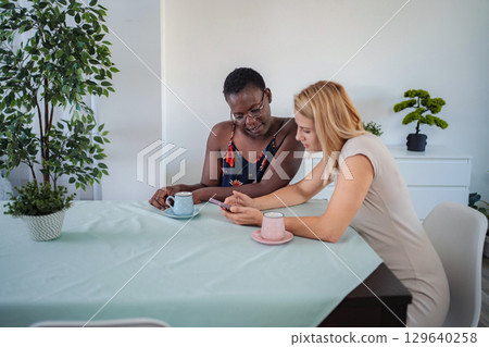 Two Young Women Looking at Smartphone and Drinking Tea at Home 129640258