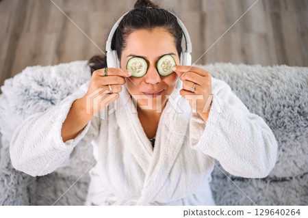 Woman applying cucumber slices on eyes while listening to music and wearing bathrobe Woman applying cucumber slices on eyes while listening to music and wearing bathrobe 129640264