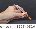 Close-Up View of a Hand Holding a Medical Syringe with an Orange Tip against a Dark Background for Exploration of Health and Medical Topics 129640310