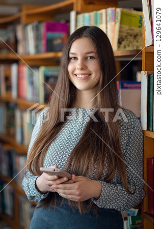 Portrait of an attractive brunette young girl standing with phone in her hands in the library. Intelligent brunette female student studying with pleasure 129641079