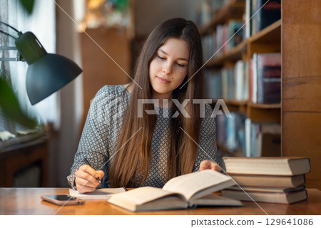 Concentrated young female student sitting at the table, reading and writing something for her studying. Young brunette girl working at the library, doing her hometasks Concentrated young female student sitting at the table, reading and writing something for her studying. Young brunette girl working at the library, doing her hometasks 129641086