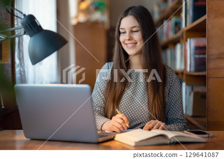 Cute young student girl sitting at a table with laptop in front of her, writing something in the notebook. Concept of education, studying in a library, modern students 129641087