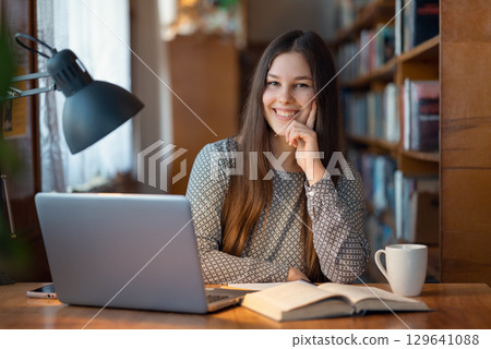 Cheerful female student sitting at a table in library with laptop, book and cup of tea in front of her. Enjoying students life, getting knowledge with pleasure Cheerful female student sitting at a table in library with laptop, book and cup of tea in front of her. Enjoying students life, getting knowledge with pleasure 129641088