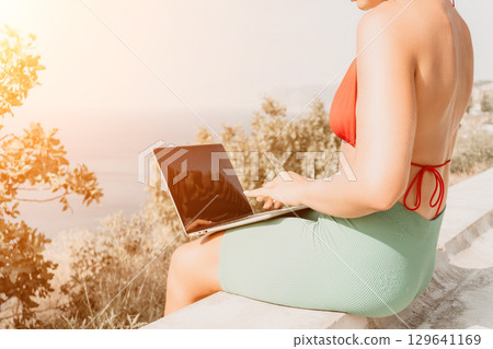 Woman Laptop Beach Work - Woman works on a laptop while sitting on a stone wall overlooking a beach. Woman Laptop Beach Work - Woman works on a laptop while sitting on a stone wall overlooking a beach. 129641169