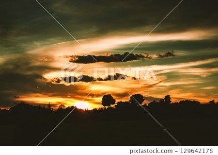 A stunning sunset paints the sky with vibrant orange gold hues, a dramatic, serene landscape scene. The sun dips low behind the silhouetted trees. A stunning sunset paints the sky with vibrant orange gold hues, a dramatic, serene landscape scene. The sun dips low behind the silhouetted trees. 129642178