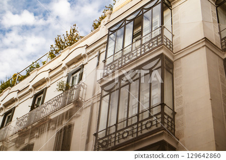 Elegant building facade with intricate balconies, windows, rooftop greenery against a sky backdrop. An architectural view of a building's exterior. 129642860