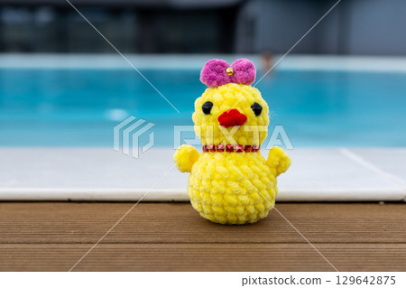 A cute, handmade crochet chick wearing a pink bow sits by a pool. Summertime vacation. A funky chicken toy against the blue water in a swimming pool. 129642875