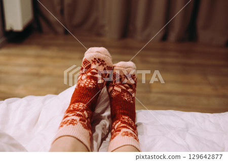 Female feet wearing festive patterned socks, relaxing on bed indoors. Cozy feet resting on sofa, warm, patterned winter socks. Comfort relaxing moment Female feet wearing festive patterned socks, relaxing on bed indoors. Cozy feet resting on sofa, warm, patterned winter socks. Comfort relaxing moment 129642877