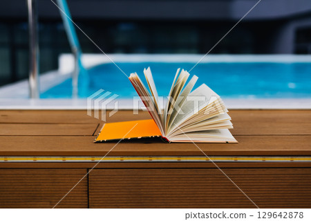 An open book sits peacefully near a pool offering a moment of relaxation A paper book rests on a wooden surface near a swimming pool Leisurely reading An open book sits peacefully near a pool offering a moment of relaxation A paper book rests on a wooden surface near a swimming pool Leisurely reading 129642878