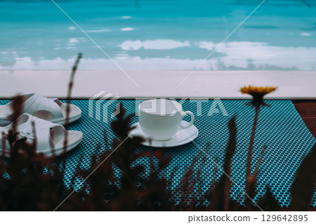 A tranquil scene showcases a cup, slippers, and flower near a pool in summer. A serene poolside scene features a drink. Calm blue water. Summer mood. 129642895