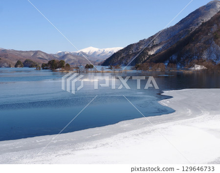 Lake Akimoto in Urabandai in winter. The partially frozen lake's blue surface is covered with white ice waves, and the snow-capped Mount Minowa stands out against the blue sky, creating a striking and beautiful sight. Lake Akimoto in Urabandai in winter. The partially frozen lake's blue surface is covered with white ice waves, and the snow-capped Mount Minowa stands out against the blue sky, creating a striking and beautiful sight. 129646733