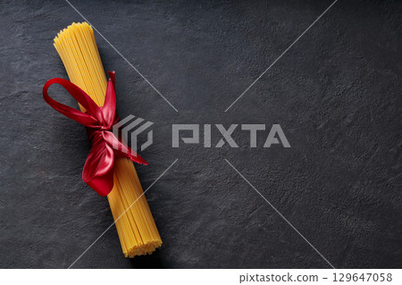 Raw Spaghetti Bundle Tied with Red Ribbon on Dark Background Top View Copy Space Raw Spaghetti Bundle Tied with Red Ribbon on Dark Background Top View Copy Space 129647058