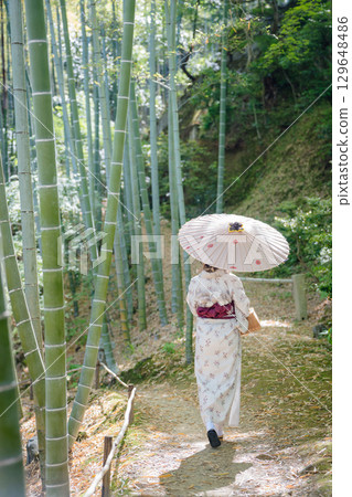 A woman in a kimono holding a Japanese umbrella strolls along a bamboo forest path 129648486
