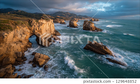 Remote windswept coastline with dramatic rock arch, rugged cliffs, ocean waves, moody sky 129648852