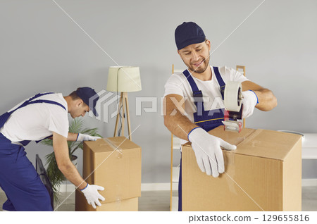 Two professional young male movers securing cardboard boxes with adhesive tape in new home. 129655816
