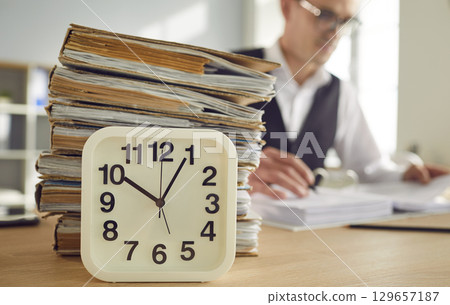 Overworked business man with the clock at his workplace and with a pile of folders and documents. 129657187