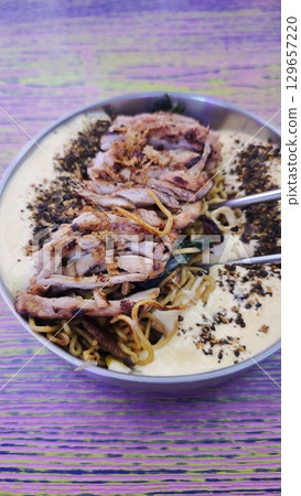 Grilled chicken served over creamy ramen noodles with spices and vegetables in a metal bowl. The image represents fusion cuisine, comfort food, and a bold flavorful dining experience 129657220