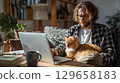 Man working on laptop at home with fluffy ginger cat on lap in cozy sunlight 129658183